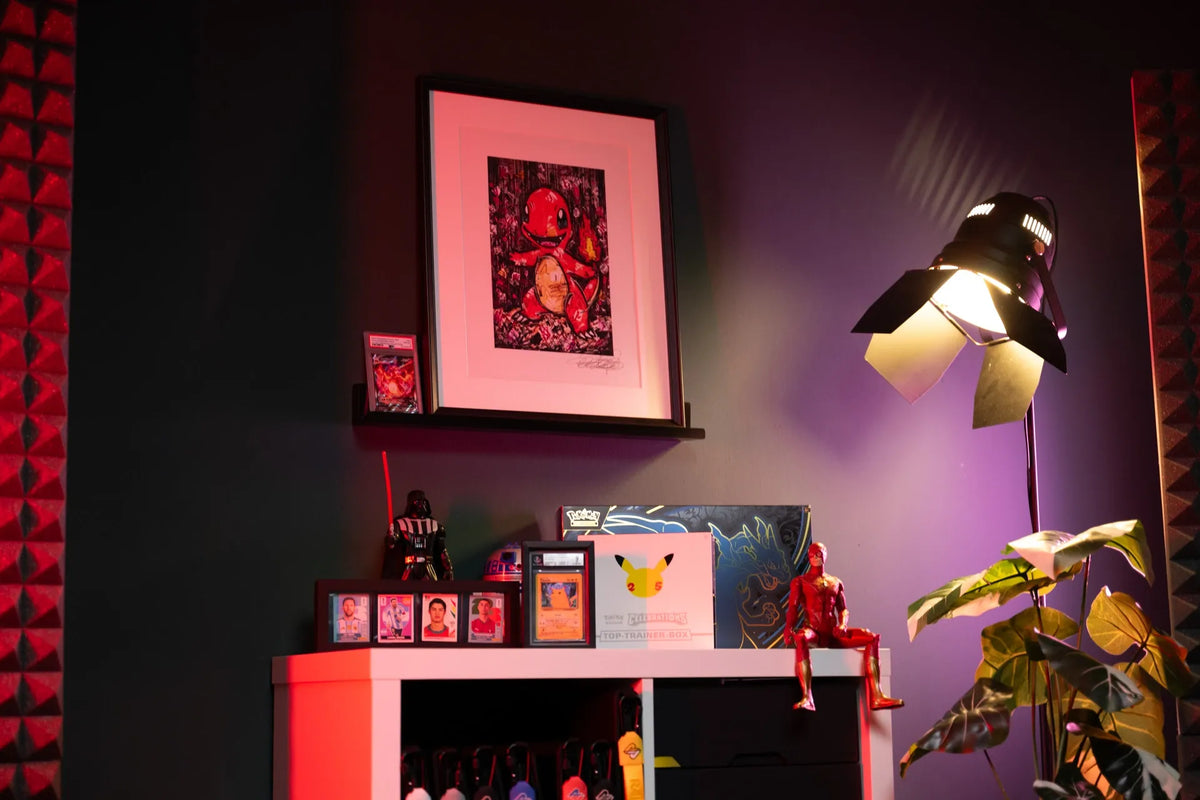 A framed art print featuring a red and black illustration of a character, displayed on a shelf in a styled room with ambient red and purple lighting, a studio spotlight, and a potted plant.
